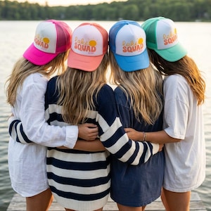 Puede incluir: Cuatro mujeres con gorras de camionero coloridas con el texto "Lake Squad" y un gr&aacute;fico de sol. Las gorras son rosas, coral, azules y verdes. Est&aacute;n de pie en un muelle de madera, abraz&aacute;ndose.