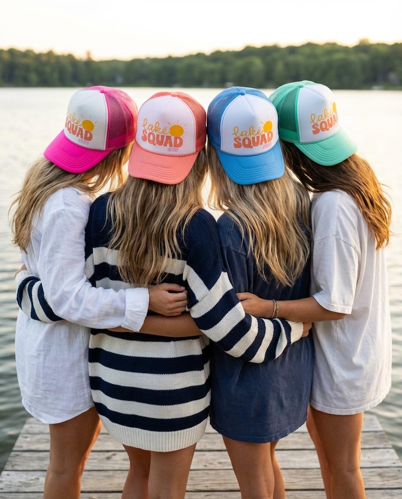 Puede incluir: Cuatro mujeres con gorras de camionero coloridas con el texto "Lake Squad" y un gr&aacute;fico de sol. Las gorras son rosas, coral, azules y verdes. Est&aacute;n de pie en un muelle de madera, abraz&aacute;ndose.