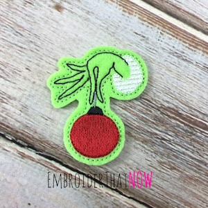 May include: A green felt Christmas ornament applique with a red ball and black outline. The applique features a hand reaching for the ornament. The text "EMBROIDER THAT NOW" is printed at the bottom.