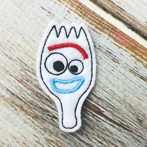 Pode incluir: Um personagem branco em forma de garfo de feltro com um rosto sorridente. O personagem tem olhos pretos grandes, uma sobrancelha vermelha e uma boca azul. O contorno é preto, em um fundo de madeira.