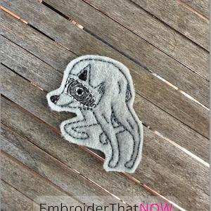 May include: A light gray felt patch featuring a black embroidered outline of a raccoon with a distinctive black eye mask and pointed ears. The patch is shaped like the animal and includes the text EmbroiderThatNOW at the bottom.