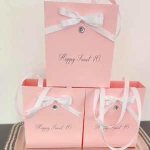 Puede incluir: Bolsas de regalo rosas con lazos de cinta blanca y un adorno de pedrería. Las bolsas tienen asas y el texto "Happy Sweet 16" impreso en ellas. Las bolsas están apiladas en una bandeja plateada.
