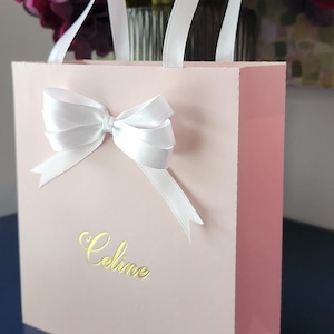 Könnte beinhalten: Eine rosa Geschenkverpackung mit weißen Schleifenhenkeln und einer Schleife. Das Wort "Celine" steht in goldener Schrift auf der Vorderseite. Dunkelrote Blumen sind im Hintergrund.