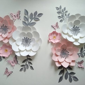 Pu&ograve; includere: Un set di decorazioni murali con fiori di carta rosa e bianchi con foglie grigie e farfalle. I fiori sono sovrapposti e hanno un effetto 3D.