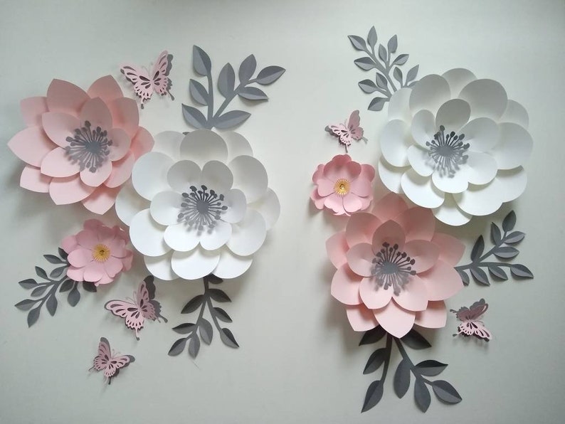 Pu&ograve; includere: Un set di decorazioni murali con fiori di carta rosa e bianchi con foglie grigie e farfalle. I fiori sono sovrapposti e hanno un effetto 3D.