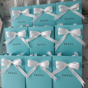 May include: Nine turquoise blue gift bags with white satin ribbon bows and the text "Liz & Co." printed on the front.