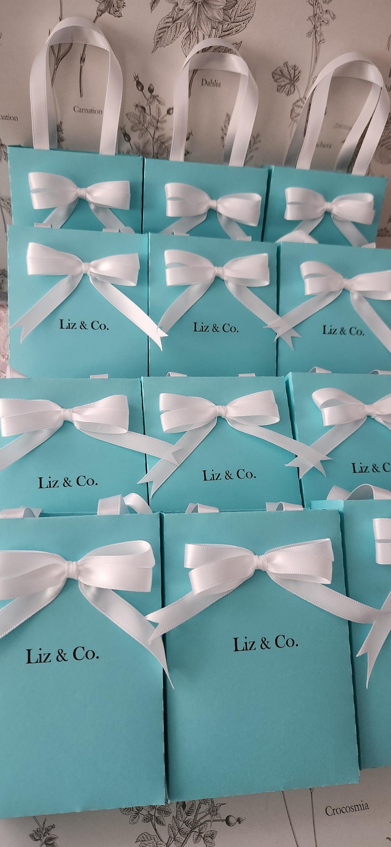 May include: A group of 12 turquoise blue gift boxes with white satin ribbon bows and the text "Liz & Co." printed on the front of each box.