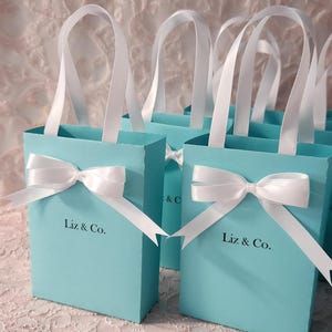 May include: Set of small turquoise blue gift bags with white satin ribbon handles and bows. Each bag has the text "Liz & Co." printed on the front.