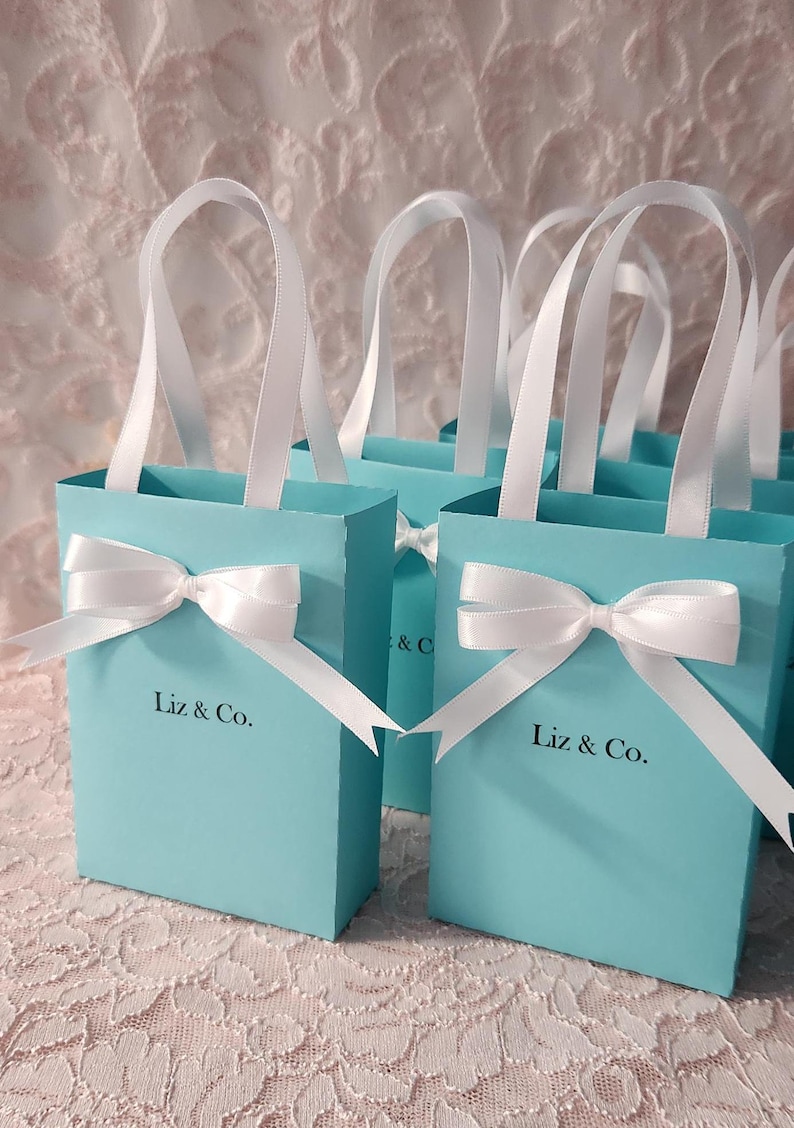 May include: Set of small turquoise blue gift bags with white satin ribbon handles and bows. Each bag has the text "Liz & Co." printed on the front.