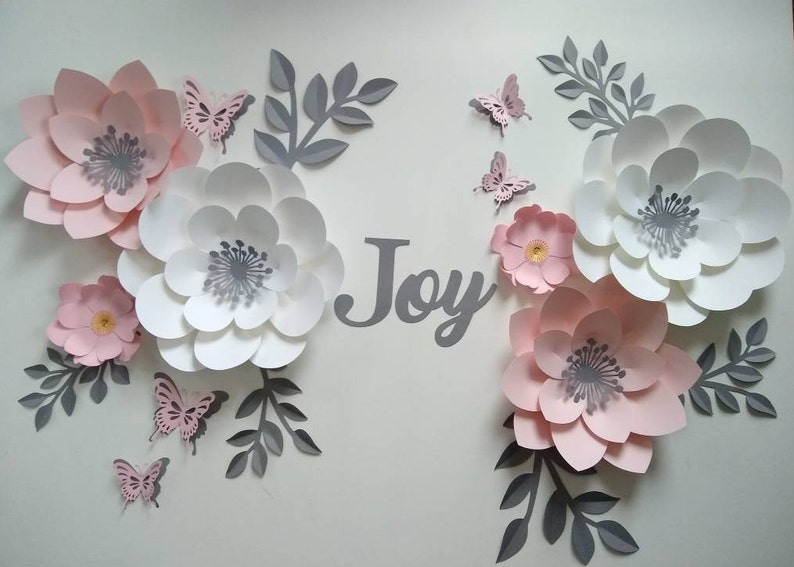 Pu&ograve; includere: Un ritaglio di carta grigia della parola "Joy" circondato da fiori di carta rosa e bianchi e foglie di carta grigia. Ci sono anche diverse piccole farfalle di carta rosa.