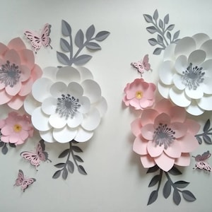 Pu&ograve; includere: Un set di fiori di carta rosa e bianchi con foglie grigie e farfalle. I fiori sono sovrapposti e hanno un effetto 3D.