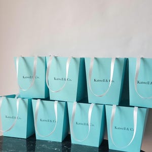 May include: Eight turquoise blue gift boxes with white ribbon handles and the text "Katwell & Co." printed on the front.