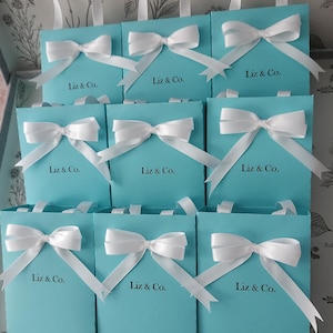 May include: Nine turquoise blue gift bags with white ribbon bows and the text "Liz & Co." printed on the front.