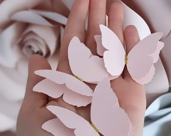 May include: Four delicate pink paper butterflies with gold glitter accents. The butterflies are held in a hand against a blurred background.