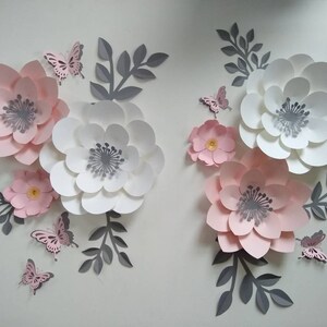 Pu&ograve; includere: Un set di fiori di carta rosa e bianchi con centri grigi. I fiori sono disposti in un motivo decorativo con farfalle e foglie.