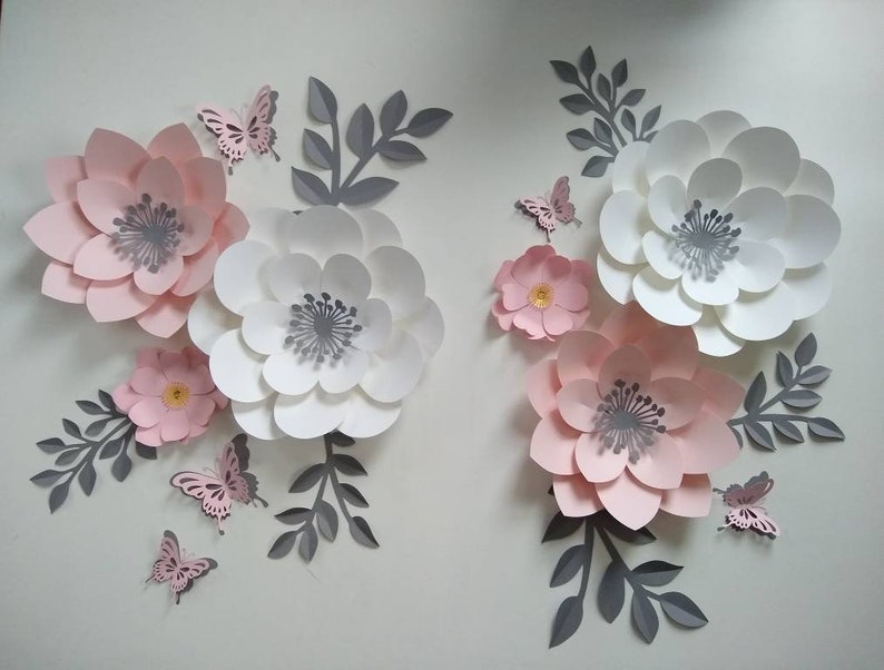 Pu&ograve; includere: Un set di fiori di carta rosa e bianchi con centri grigi. I fiori sono disposti in un motivo decorativo con farfalle e foglie.