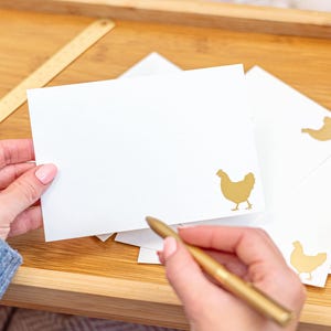 May include: A white stationery set featuring gold chicken designs. The set includes blank cards and envelopes. A gold pen is held over a card, suggesting writing. The items are arranged on a wooden tray with a candle and decorative matches.