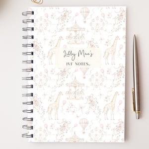 May include: A spiral-bound notebook with a white cover featuring a repeating pattern of giraffes, carousels, hot air balloons, and flowers in soft pink and brown tones. The centre text reads Lilly Mae's IVF NOTES..