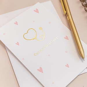 May include: A greeting card with a gold stethoscope heart design and the word "Congratulations!" is displayed on a light pink surface. The card is decorated with small pink hearts. A gold pen, a gold binder clip, and a spiral notebook are also visible.
