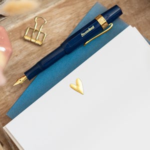 May include: A navy blue fountain pen with gold detailing lies on a blue notebook and white paper. A gold heart-shaped sticker and a gold binder clip are also present. A small pink vase with dried flowers is in the background.