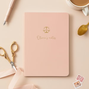 May include: A light pink notebook with rounded corners featuring a gold foil scale of justice icon and the text Olivia's notes printed in a cursive font on the front cover. The notebook is positioned centrally with a pen, scissors, and stamps.