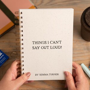 May include: A spiral-bound notebook with the text "THINGS I CAN'T SAY OUT LOUD!" is held by hands. A brown travel mug and stationery items are also visible. The notebook has a speckled, off-white cover and gold spiral binding.