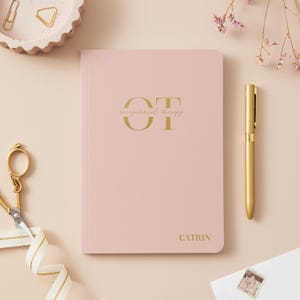 May include: A dusty pink notebook with gold foil lettering that reads OT occupational therapy and the name CATRIN at the bottom. The notebook is accompanied by a gold pen, gold scissors with a white ribbon, and a small white envelope with a stamp.