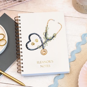 May include: A cream-coloured spiral-bound notebook. The cover displays a watercolour illustration of a stethoscope shaped like a heart, with paw prints inside. The text "ELEANOR'S NOTES" is printed below the stethoscope in gold lettering.