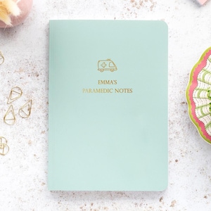 May include: A light mint green notebook with gold foil lettering and a gold ambulance icon. The text on the cover reads EMMA'S PARAMEDIC NOTES. The notebook has a smooth, matte finish and rounded corners, presenting a clean and professional appearance.