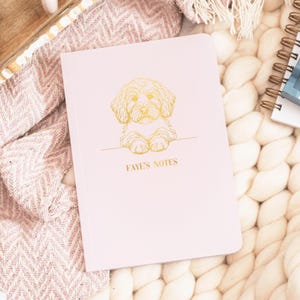 May include: A pale pink notebook featuring a gold outline of a dog and the words "FAYE'S NOTES". The notebook rests on a white, textured surface, next to a pink and white patterned blanket.