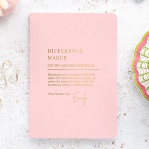 May include: A pink notebook with gold text reading "DIFFERENCE MAKER" and a definition. The name "Emily" is also printed on the notebook. The notebook is on a white surface with decorative items around it.