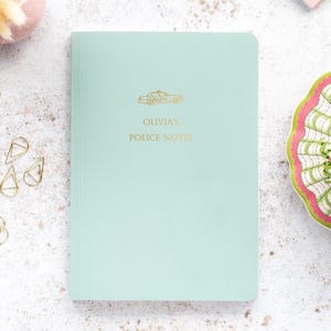 May include: A pale teal notebook featuring gold lettering and a police car illustration. The text reads "Olivia's Police Notes". The notebook is closed and has a smooth cover. The notebook is on a light-coloured surface.