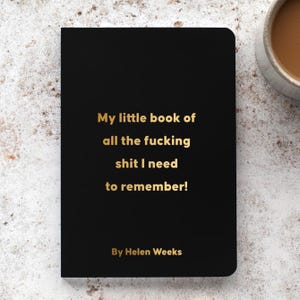 May include: Black notebook with gold lettering that reads "My little book of all the fucking shit I need to remember!" by Helen Weeks. The notebook is on a light-colored surface.