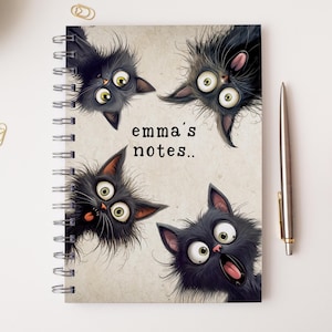 May include: A notebook with a spiral binding, decorated with a cartoon design of four black cats with large eyes. The cover includes the text "emma's notes..". A silver pen is positioned to the right.