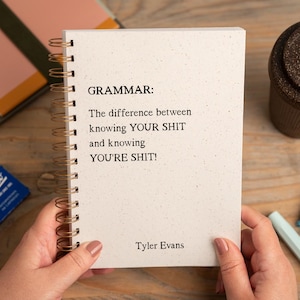 May include: A cream-coloured spiral-bound notebook with the text "GRAMMAR: The difference between knowing YOUR SHIT and knowing YOU'RE SHIT!" is held by hands. A brown travel mug, pens, and Kaweco pen boxes are also visible.