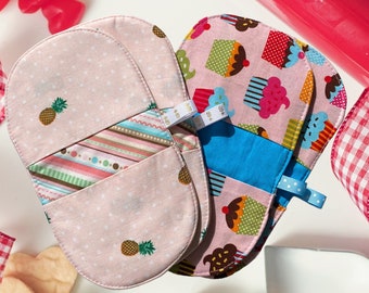 May include: Two oven mitts with a pink polka dot and pineapple pattern, and a cupcake pattern. The mitts have blue and white ribbon loops. Heart-shaped cookie cutters and candies are also visible.