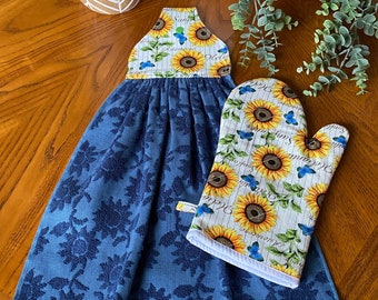 May include: A blue floral kitchen towel with a sunflower and butterfly print hanging from a loop. A matching oven mitt with the same sunflower and butterfly pattern is next to the towel. The towel and mitt are on a wooden surface.