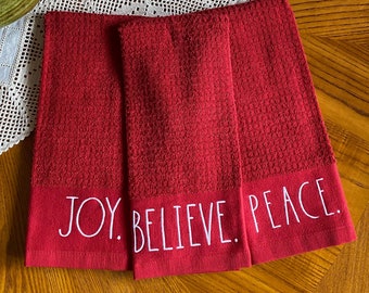 May include: Three red hand towels with the words "JOY. BELIEVE. PEACE." in white lettering. The towels are made of a textured fabric and are folded neatly. The towels are on a wooden surface.