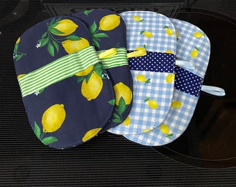May include: Four oven mitts with lemon patterns. Two mitts have a navy blue background with yellow lemons and green leaves. The other two mitts have a blue and white gingham and a blue polka dot pattern. A sign that says "KITCHEN CLOSED" is in the background.