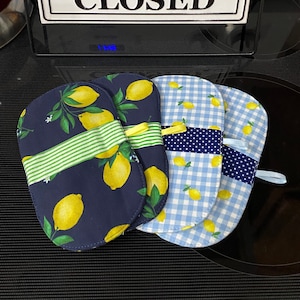 May include: Four oven mitts with lemon patterns. Two mitts have a navy blue background with yellow lemons and green leaves. The other two mitts have a blue and white gingham and a blue polka dot pattern. A sign that says "KITCHEN CLOSED" is in the background.