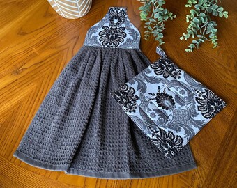 May include: A gray kitchen towel with a decorative top featuring a black and white floral pattern. A matching pot holder with the same pattern is next to the towel. The towel is hanging on a wooden surface.