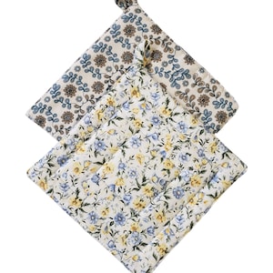 May include: Two quilted pot holders with floral patterns. One features blue and brown flowers on a cream background, and the other has blue and yellow flowers on a white background. Each has a loop for hanging.
