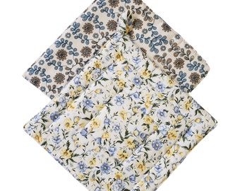 May include: Two quilted pot holders with floral patterns. One features blue and brown flowers on a cream background, and the other has blue and yellow flowers on a white background. Each has a loop for hanging.