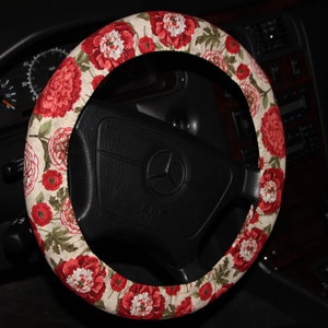 May include: A steering wheel cover with a floral design. The cover has a pattern of red and white flowers with green leaves on a beige background. The cover is fitted on a black steering wheel.