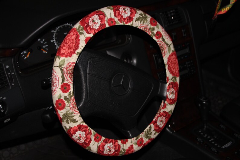 May include: A steering wheel cover with a floral design. The cover has a pattern of red and white flowers with green leaves on a beige background. The cover is fitted on a black steering wheel.