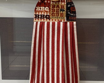 May include: A hanging kitchen towel with a red and white striped bottom and a patterned top. The top features the words "BBQ" and "Barbeque Forever" with images of grilling tools and a pig. The towel is attached to a white oven.