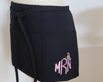 May include: Black waist apron with two pockets and tie closure. The apron features embroidered initials "MRN" in pink, accompanied by a colorful butterfly design. The apron is displayed on a mannequin.