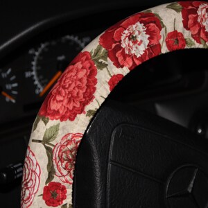 May include: A floral steering wheel cover with a pattern of large red and white peonies and smaller red flowers on a cream background. The cover is fitted on a black steering wheel, with a car dashboard visible in the background.