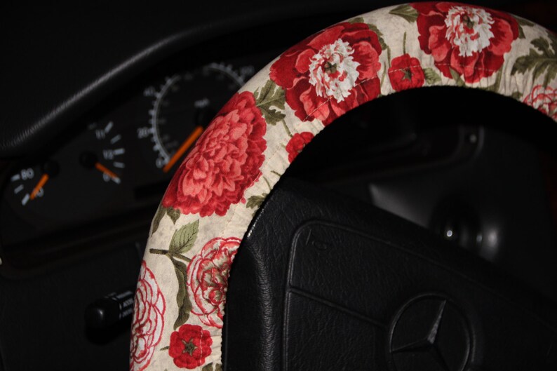 May include: A floral steering wheel cover with a pattern of large red and white peonies and smaller red flowers on a cream background. The cover is fitted on a black steering wheel, with a car dashboard visible in the background.