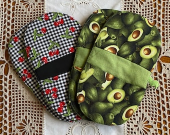 May include: Two oven mitts. One mitt features a black and white checkered pattern with red cherries. The other mitt has a green avocado print. Both mitts have a black or green band for the hand.